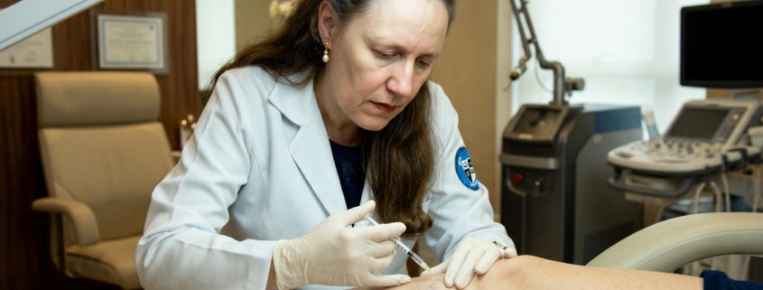 A dermatologista Prof. Dra. Marcia Ferraz Nogueira, vestindo jaleco clínico branco e luvas estéreis, trabalha concentrada ao aplicar uma injeção de escleroterapia em vasinhos azulados na perna de uma paciente. A cena é iluminada por uma lâmpada cirúrgica em um consultório dermatológico sofisticado em São Paulo, com aparelhos médicos de alta tecnologia ao fundo.